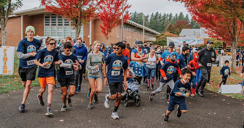 Runners of all ages running from the starting line at the Superhero 5K surrounded by trees with red and orange fall leaves.