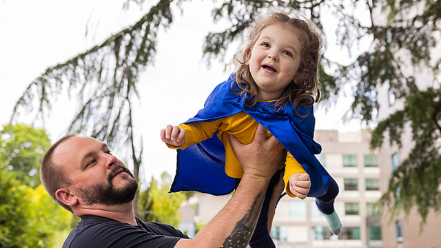 Little girl in yellow shirt and blue cape is lifted in air by her father
