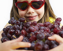 A close photo of a school aged girl wearing red heart-shaped sunglasses and holding a large bunch of purple grapes