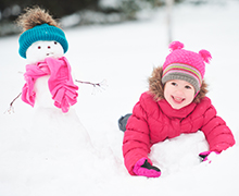 A preschool aged girl wearing a pink jacket and hat while laying in the snow next to a snowman