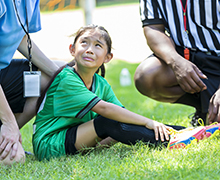 Young girl in a soccer uniform is clutching her leg while sitting on the ground