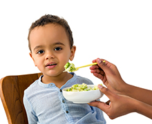 A toddler boy sitting in a high-chair turns away from food that is being fed to him