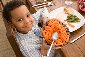 A little boy sitting at the dinner table holding up a big bowl of carrots with a spoon in it