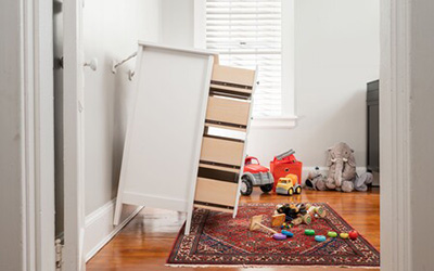 A tall, light-wood dresser has tipped forward on a rug in a bright room, showcasing the danger of unsecured furniture. Toys are scattered nearby.