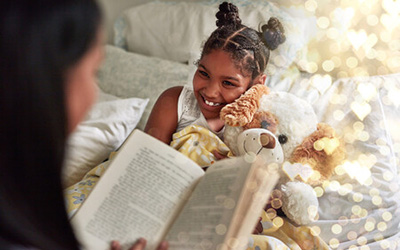 A joyful young girl with braided pigtails lies comfortably in bed with a stuffed animal, smiling brightly while an adult reads a book to her. The scene is lit with warm, magical-looking bokeh lights.