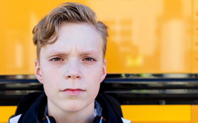 A close-up portrait of a blonde young person with a worried or determined expression, wearing headphones around their neck, with the bright yellow side of a school bus behind them.