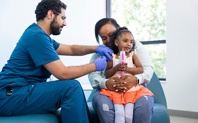 A male healthcare professional in blue scrubs and gloves prepares to give a young girl an injection while she is held securely and comforted by an adult woman.