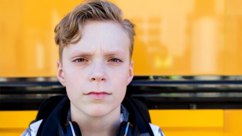 A close-up portrait of a blonde young person with a worried or determined expression, wearing headphones around their neck, with the bright yellow side of a school bus behind them.