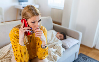 A parent calls a doctor or helpline about her sick child resting on the sofa.
