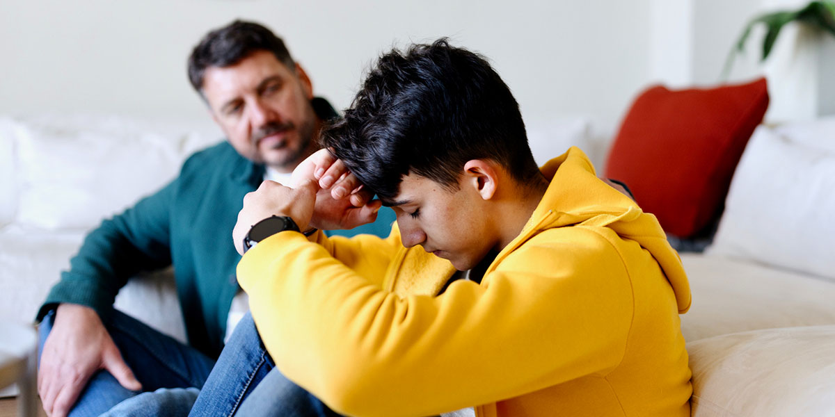A boy sits with his head in his hands as his father looks on