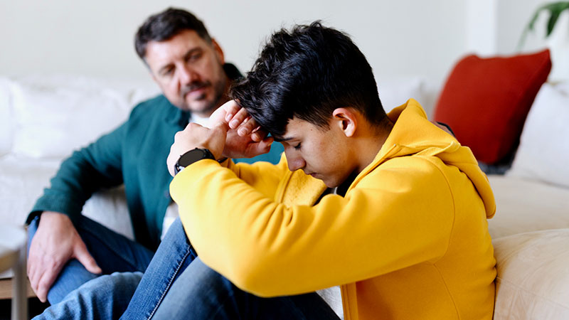 A boy holds his head in his hands as his father looks on
