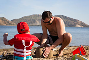 A man and a toddler in a life jacket build sand castles.