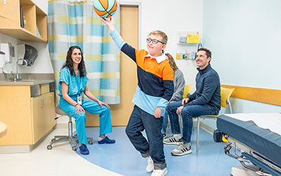 A boy tosses a basketball in a doctor's office while his parents and doctor look on