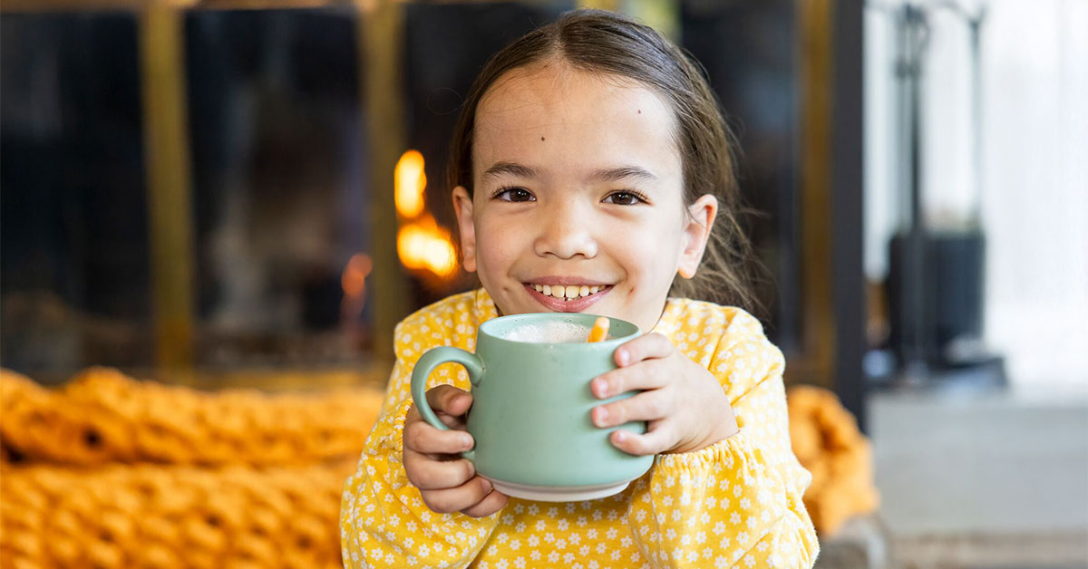 Smiling girl holds blue mug of cocoa in front a fireplace