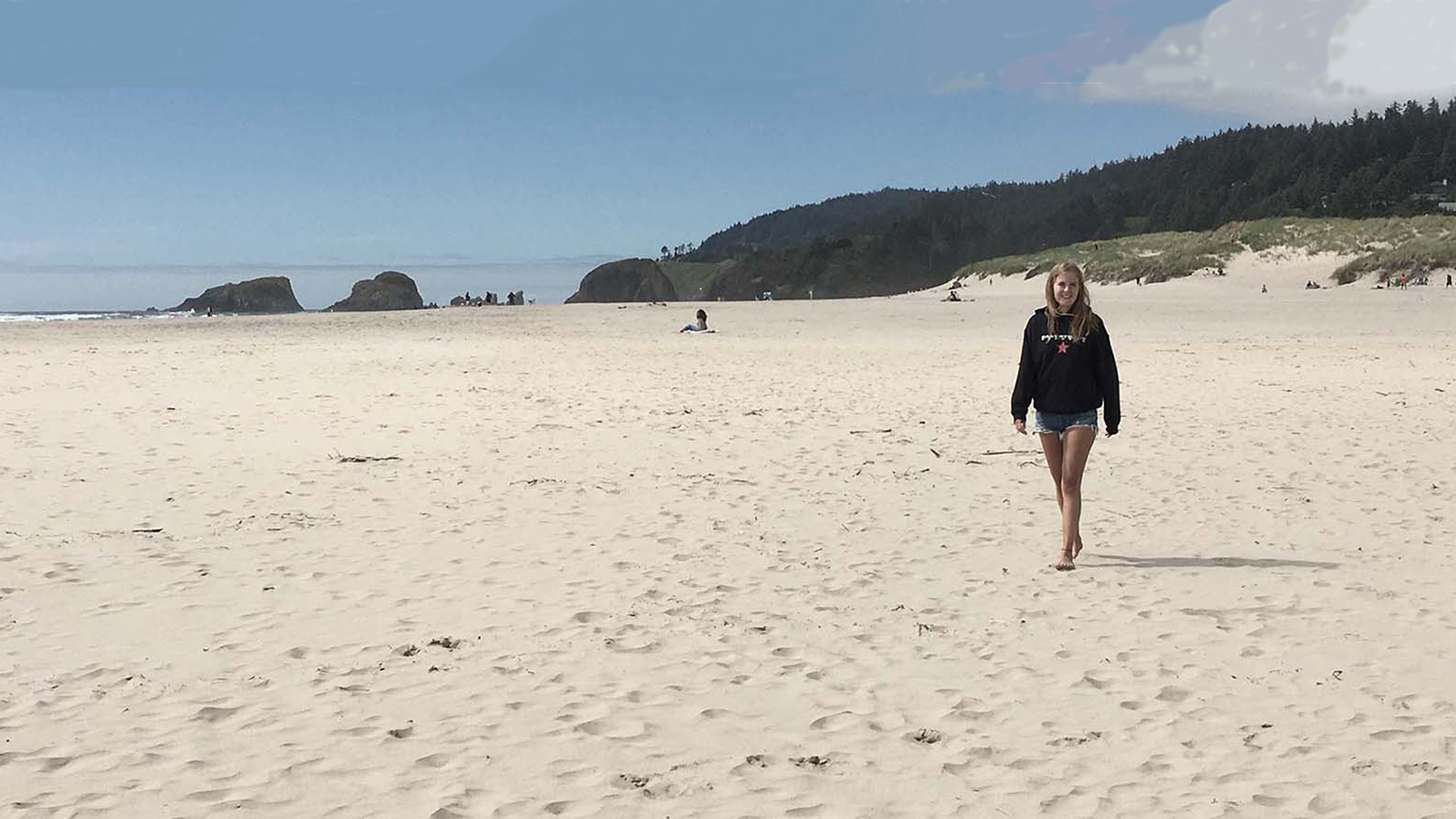 A teen girl wearing jean shorts and a black hoodie smiles as she walks on a Pacific Northwest beach.
