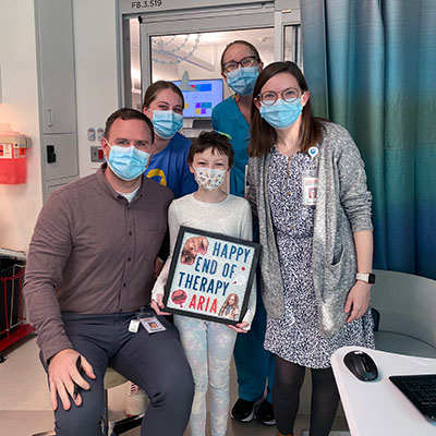 A hospital photo - A girl holds a sign that reads "Happy end of therapy, Aria". She is surrounded by healthcare providers