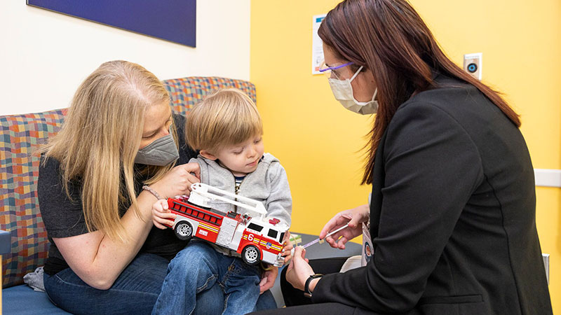 A boy with a firetruck sites in his mom's lap while receiving a shot from a Seattle Children's provider