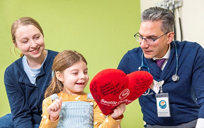 A girl holds a heart-shaped pillow as her mother and doctor look on