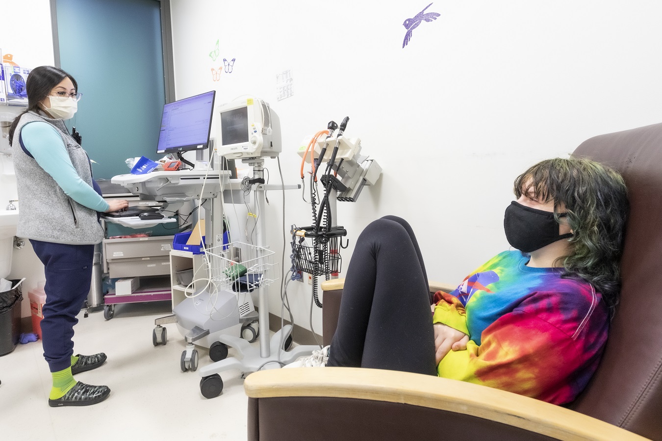 A nurse speaks with a seated patient in a tie-dyed sweatshirt