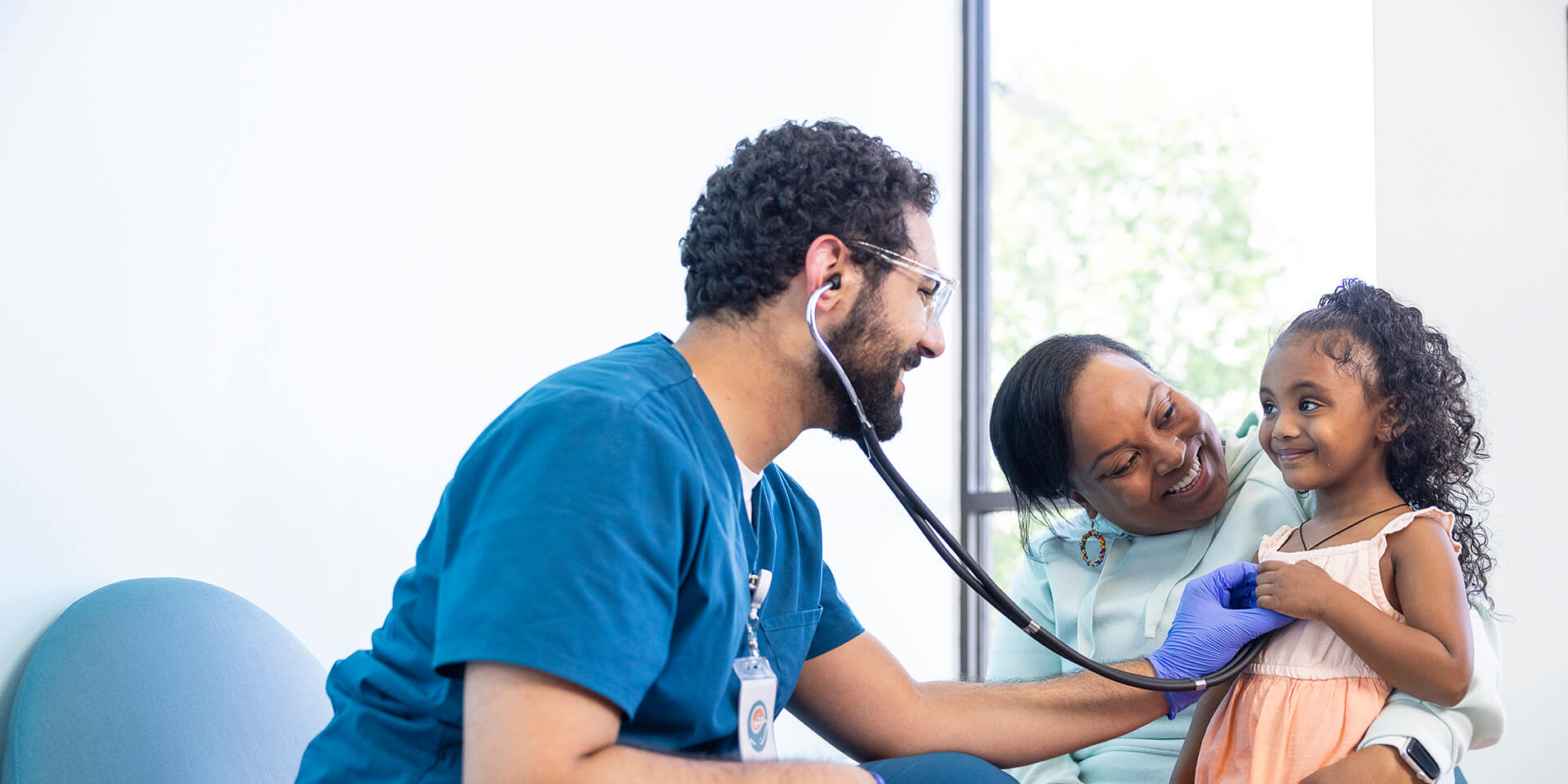 A provider listens to a smiling young girl's heart with a stethoscope while her mother watches