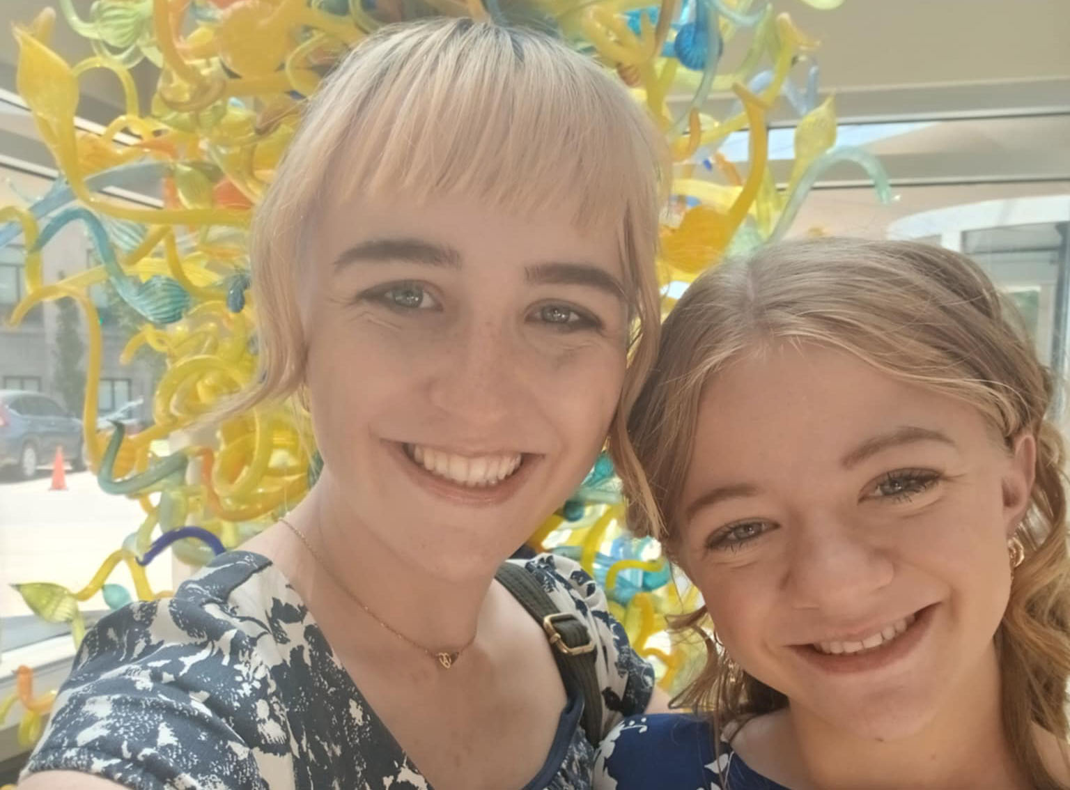 Two smiling sisters, Mary (left) and Audrey (right), pose for a close-up photo in front of a large, bright yellow and orange glass sculpture.