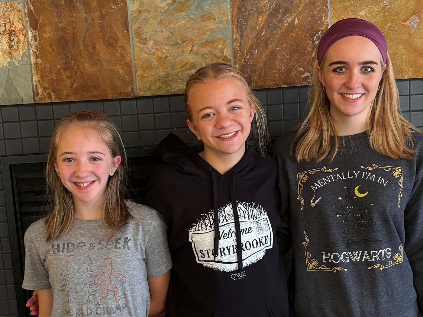 Three sisters smiling in casual clothing: transplant recipient Mary (right) and donor Audrey (center) pose with their younger sister (left) in front of a stone wall.