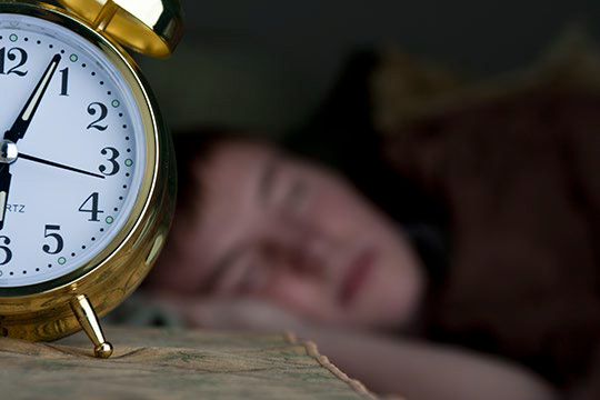 An alarm clocks sits in the foreground while a teen sleeps in the background