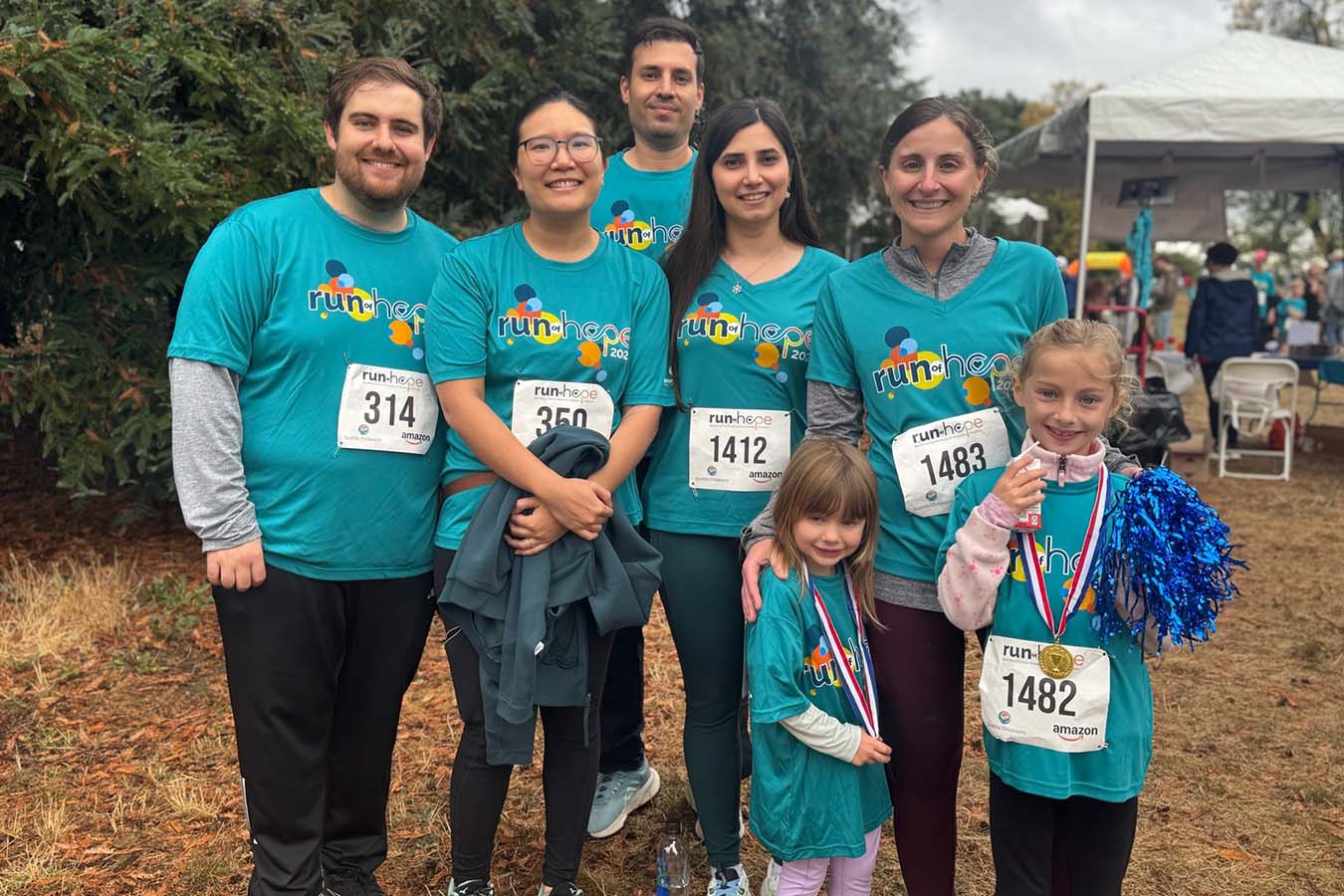 A photograph of a group of eight people, all wearing matching teal "run-hope" t-shirts with bib numbers, gathered outside on a grassy area. The participants include men, women, and two young girls, one of whom is holding a medal and a blue pom-pom.
