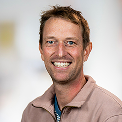 Sylvain Simon, PhD, a fair-skinned man with blue eyes and short, sandy-brown hair, smiling at the camera against a blurred, light-colored background. He is wearing a light mauve or tan zip-up sweatshirt over a blue and white striped shirt.
