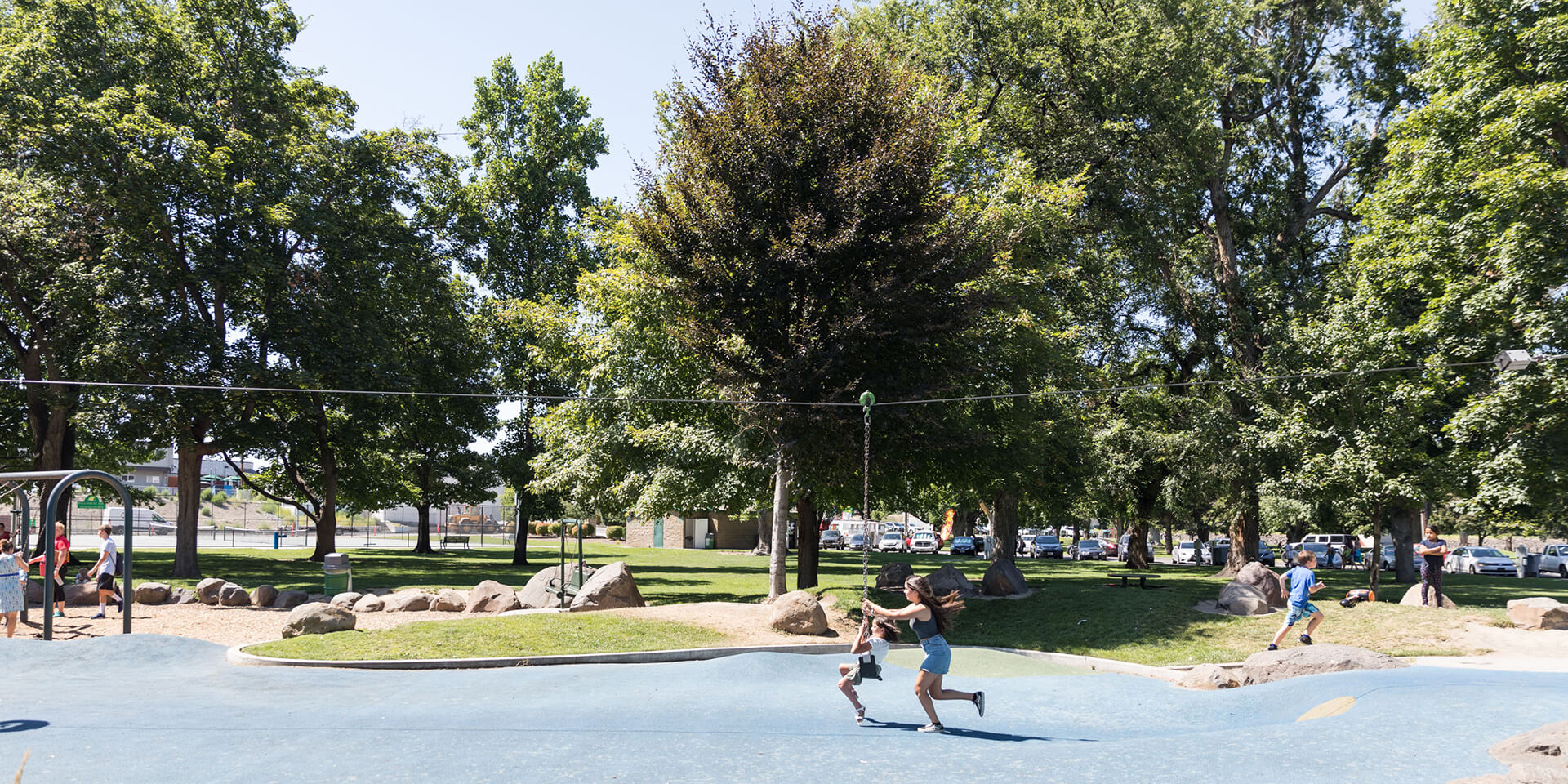 Children and adults playing in a large, sunny park playground. The blue, wavy play surface is surrounded by grassy hills and many large, mature trees. People are visible on the play surface and the surrounding grassy areas, with cars parked in the distance.