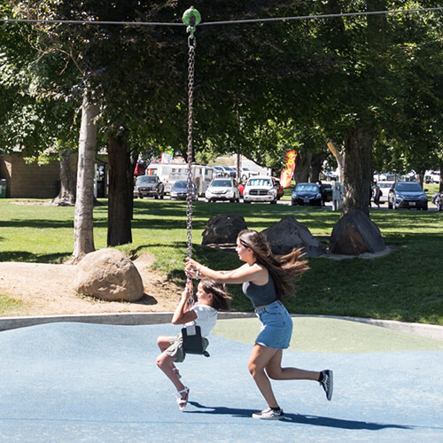 A close-up of an older girl pushing a younger girl on a zip line in a sunny park. Both girls are actively playing and smiling, with lush green trees and parked cars visible in the background.