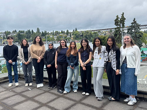 A diverse group of students stand side by side outdoors on a terrace, with trees, buildings, and an overcast sky in the background.