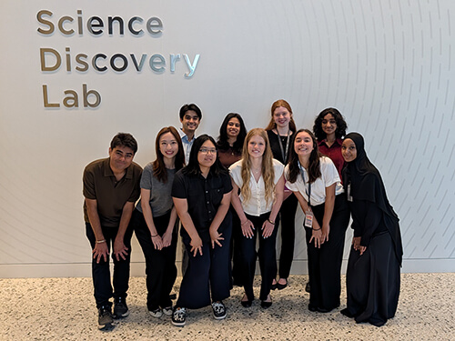 A diverse group of students stand side by side outdoors on a terrace, with trees, buildings, and an overcast sky in the background.