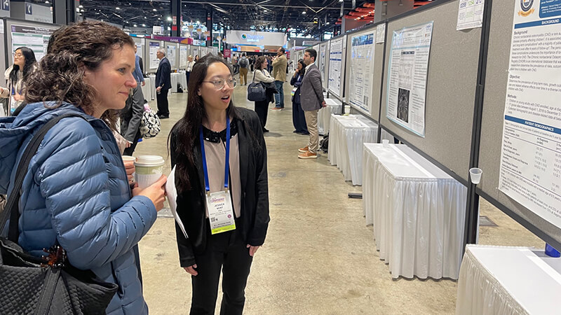 Two female conference attendees standing in an exhibition hall and discussing a large research poster display. Both are wearing lanyards, and one holds a cup and papers,