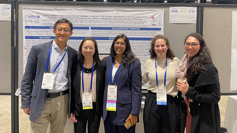 A group of five smiling researchers and presenters, three women and two men, standing together in front of their large scientific poster at a conference. The poster title is about the development of a clinically important difference measure for children with Chronic Nonbacterial Osteomyelitis.