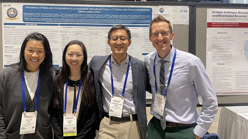 Four smiling Seattle Children's team members, two men and two women, wearing conference lanyards, standing proudly in front of their research poster titled "Prevalence of Risks and Complications in a Longitudinal International Cohort of Children with Chronic Nonbacterial Osteomyelitis (CNO)."