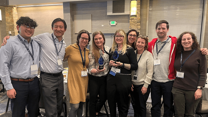 A diverse group of nine smiling people standing together indoors, wearing conference lanyards. Two of the women in the center are holding matching blue glass award trophies.