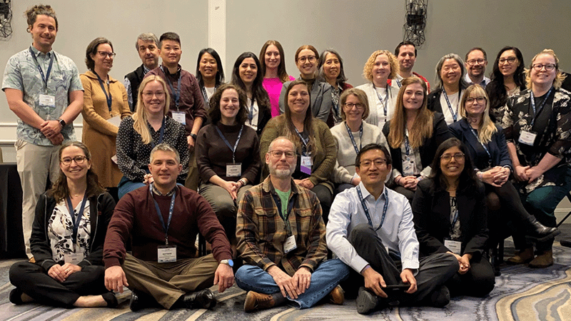 A large group photo of diverse conference attendees. The group is seated and standing in three rows, smiling at the camera and wearing lanyards.
