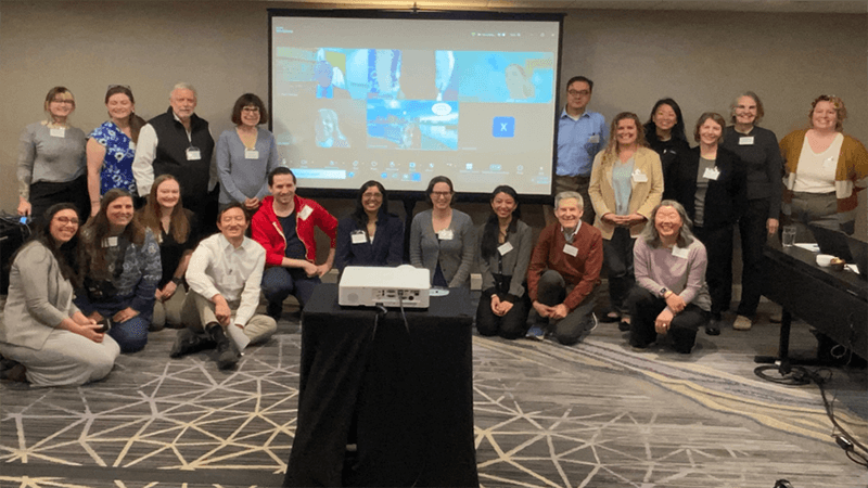 A large group of people posing in a conference room with a large projector screen behind them. The group is seated and standing, smiling at the camera.