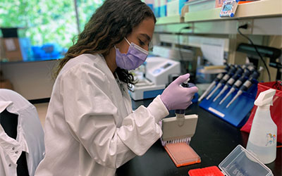 A gloved and masked lab assistant pipettes samples in a laboratory