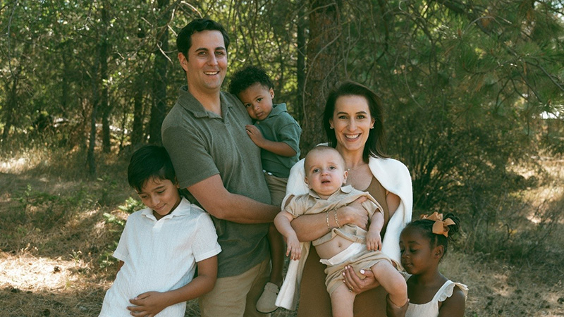 Smiling family of six poses in portrait in a forest, wearing earth toned outfits.