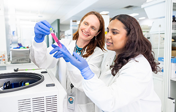 Two female researchers, Juliane Gust (left) and Mahashweta Bose (right), both wearing lab coats and blue gloves, smile while looking closely at a test tube containing a pink liquid next to a centrifuge machine.