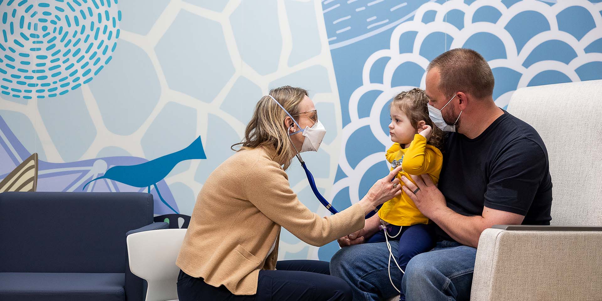 Dr. Kasey Leger listens with a stethoscope to a young patient’s heart. The patient is seated on her father’s lap.