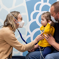 Dr. Kasey Leger listens with a stethoscope to a young patient’s heart. The patient is seated on her father’s lap.
