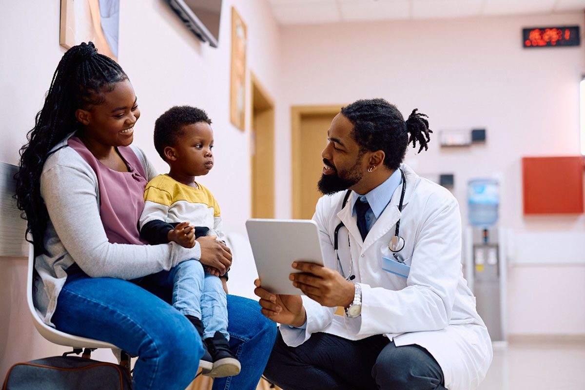 A healthcare provider in a white coat kneels to speak at eye level with a young child and their caregiver