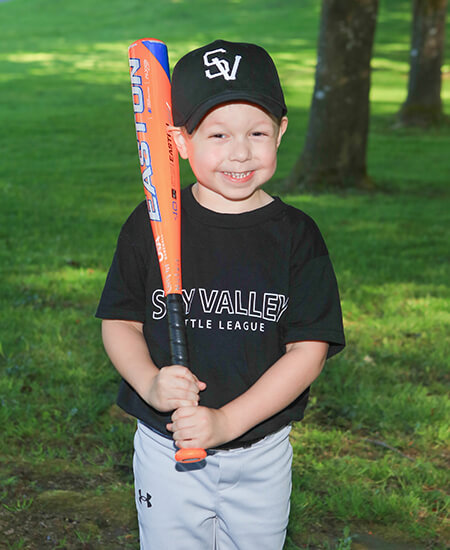 A young boy in a baseball uniform and cap holding a bat and smiling