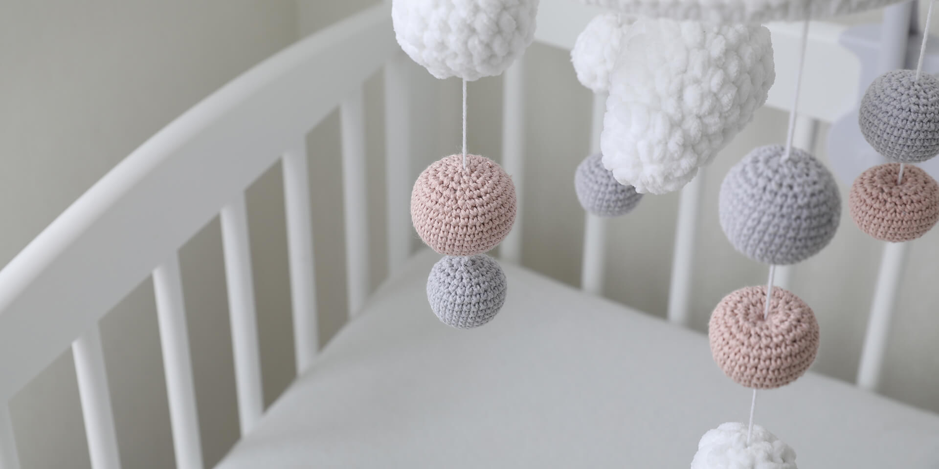 Empty, white crib with a mobile of white, grey and beige crocheted balls suspended above
